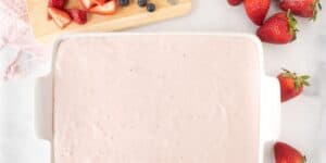 A white ceramic dish filled with pink creamy dessert is on a white surface. Next to it, a wooden cutting board holds sliced strawberries and whole blueberries. Whole strawberries are placed on the right, and a pink kitchen towel is partially visible on the left.
