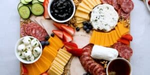 A charcuterie board featuring a variety of cheese slices, salami, fresh cucumber slices, strawberries, blueberries, and small bowls of mozzarella balls, jam, and honey. The board is artfully arranged on a light-colored surface.