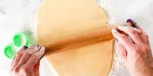 A person with purple painted nails uses a wooden rolling pin to roll out dough on a floured surface. A green heart-shaped cookie cutter is on the left side of the image. Flour is scattered around the dough and on the person's hands.