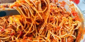 A large pot filled with spaghetti and meat sauce is being mixed with a spatula. The sauce is a rich red and chunky, containing tomato bits and ground meat. The image showcases a close-up view of the pasta dish, highlighting its texture and ingredients.