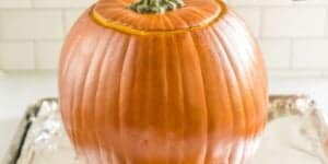 A whole pumpkin with its top cut off is placed on a foil-lined baking tray. The pumpkin has a smooth, orange skin and its hollow interior is not visible. The background consists of a white tiled wall.
