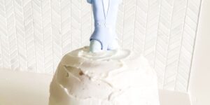 A small cake covered in white frosting, topped with a blue, plastic surfer figure. The background features a textured, white herringbone pattern on the wall.