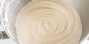 A close-up view of a metallic mixing bowl filled with smooth, creamy cake batter. The batter has a light vanilla color and is mixed into a swirl pattern in the center of the bowl. The mixing bowl is placed on a white surface.