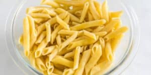 A clear glass bowl filled with perfectly cooked al dente penne pasta, coated lightly with a yellow-colored, creamy sauce. The individual pasta tubes are visibly well-defined and arranged in an appetizing manner. The background is a simple white surface.