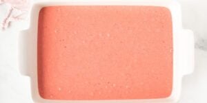 A rectangular white baking dish is filled with a smooth, pink batter. The dish is placed on a light surface, and part of a folded pink cloth is visible in the top left corner.