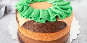 A round cake with a golden-brown base, topped with glossy chocolate frosting and decorated with a green and beige fondant ribbon arranged in a wreath pattern. The cake is placed on a scalloped-edge silver cake board, set against a light background.