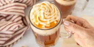 A hand holding a clear glass mug filled with a drink topped with swirled whipped cream and caramel drizzle. A striped cloth napkin is in the background.