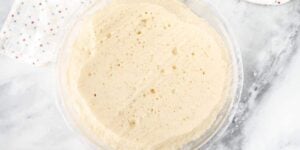 A bowl of sourdough starter sits on a marble countertop. The starter has a bubbly and slightly frothy surface, indicating active fermentation. A white cloth with small, colorful dots is partially visible in the background.
