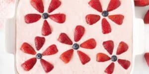 A white rectangular dish filled with pink strawberry-flavored dessert is decorated with slices of strawberries arranged in flower shapes. Each "flower" has five strawberry slices forming petals with a blueberry in the center. Whole strawberries are nearby.