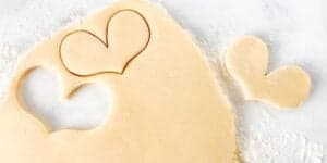 A sheet of dough is rolled out on a lightly floured surface. Heart-shaped cookie cutters are used to cut heart shapes from the dough, leaving empty heart outlines behind. Two heart-shaped dough cutouts are placed near the sheet.