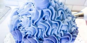 A close-up of a cake covered in blue and purple ruffled buttercream icing. A piping bag with a star tip, partially visible on the right, is being used to add more icing to the cake. The background appears to be a kitchen counter with white tiles.