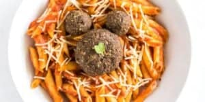 A white bowl filled with penne pasta in a red sauce, topped with grated cheese. Three round meatballs are placed on top, with a small green leaf garnish on the center meatball. The presentation resembles a face.
