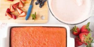 A baking scene with a rectangular strawberry cake in a white baking dish, halved strawberries and blueberries on a wooden cutting board, a blue-handled knife, a bowl of pink batter, and several whole strawberries on the side.