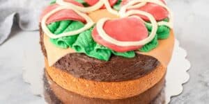 A creatively decorated cake made to look like a pizza. It features realistic-looking toppings made from frosting or fondant, including green leaves resembling spinach, red slices mimicking pepperoni, and white rings imitating onions, all atop a brown frosted layer.