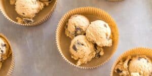 Brown paper cupcake liners filled with scoops of cookie dough containing chocolate chips are placed on a metallic surface. The image shows multiple liners, some partially visible, each containing several scoops of the dough.