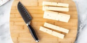 A black-handled knife and four sticks of cheese with visible seasoning are arranged on a wooden cutting board. The cutting board rests on a grayish marble surface.