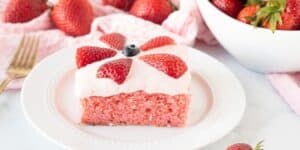 A slice of pink strawberry cake topped with white frosting, garnished with sliced strawberries and a blueberry, displayed on a white plate. Surrounding the plate are fresh strawberries, a gold fork, and a white bowl filled with strawberries on a pink cloth.