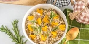 A white bowl filled with rice mixed with herbs and topped with orange flower petals is placed on a white surface. Fresh rosemary sprigs are beside the bowl, and a gold spoon rests on a green checkered cloth. Decor elements include checkered fabric pumpkins.