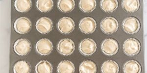 A muffin tin filled with 24 paper liners, each containing an even amount of creamy, beige batter, ready to be baked. The organized arrangement provides a neat, appealing visual of pre-baked cupcakes.