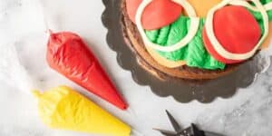 An overhead view of a cake decorated to look like a Teenage Mutant Ninja Turtle face using colorful fondant, placed on a black scalloped-edge cake board. Nearby are two piping bags filled with red and yellow icing, and a pair of black scissors.