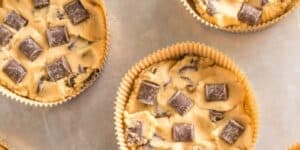 Four unbaked cookie dough muffins with chocolate chunks are placed in paper liners on a baking sheet. The dough is creamy with visible pieces of chocolate throughout each muffin.