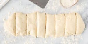 A slab of dough is rolled out on a floured surface. The dough is divided into several equal sections with a dough scraper, which is also visible in the background alongside a spoon containing more flour.