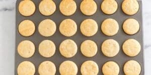 A top-down view of a baking tray filled with 24 evenly spaced, freshly baked mini cornbread muffins. The muffins are golden brown and appear uniform in size. The tray is placed on a white surface.