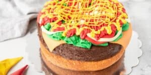 A cake decorated to look like a hamburger sits on a reflective silver board. The cake features layers mimicking a bun, patty, lettuce, cheese, tomato slices, and condiments, with colorful icing replicating ketchup, mustard, and other toppings.