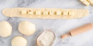 An elongated strip of raw dough is laid out with evenly spaced cubes of cheese. Next to the strip are three balls of dough, a small wooden scoop filled with flour, and a rolling pin, all set on a marble surface.