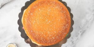 A round cake designed to look like a giant hamburger bun, complete with sesame seeds sprinkled on top. The cake sits on a dark, scalloped-edge cake board on a light gray marble surface.