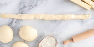 A marble countertop with three dough balls, a long rolled-out dough, a wooden rolling pin, a small bowl of flour, and cheese slices in the top right corner. The ingredients suggest preparation for baking or cooking.