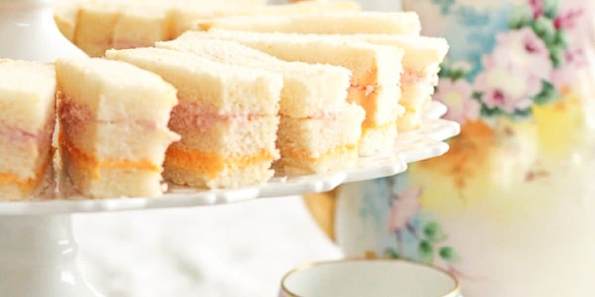 A white plate holds several neatly arranged tea sandwiches with layers of different fillings, such as jam and possibly cheese or ham. In the background, there is a floral-patterned teapot and teacup, suggesting a tea time setting.