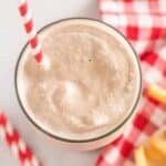 A top view of a milkshake with a red and white striped straw on a light surface. Next to the milkshake are two extra red and white striped straws and a plate of French fries placed on a red and white checkered napkin.