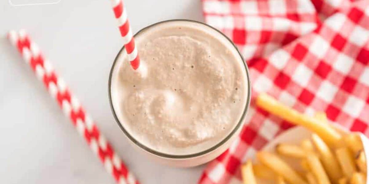 A top view of a milkshake with a red and white striped straw on a light surface. Next to the milkshake are two extra red and white striped straws and a plate of French fries placed on a red and white checkered napkin.