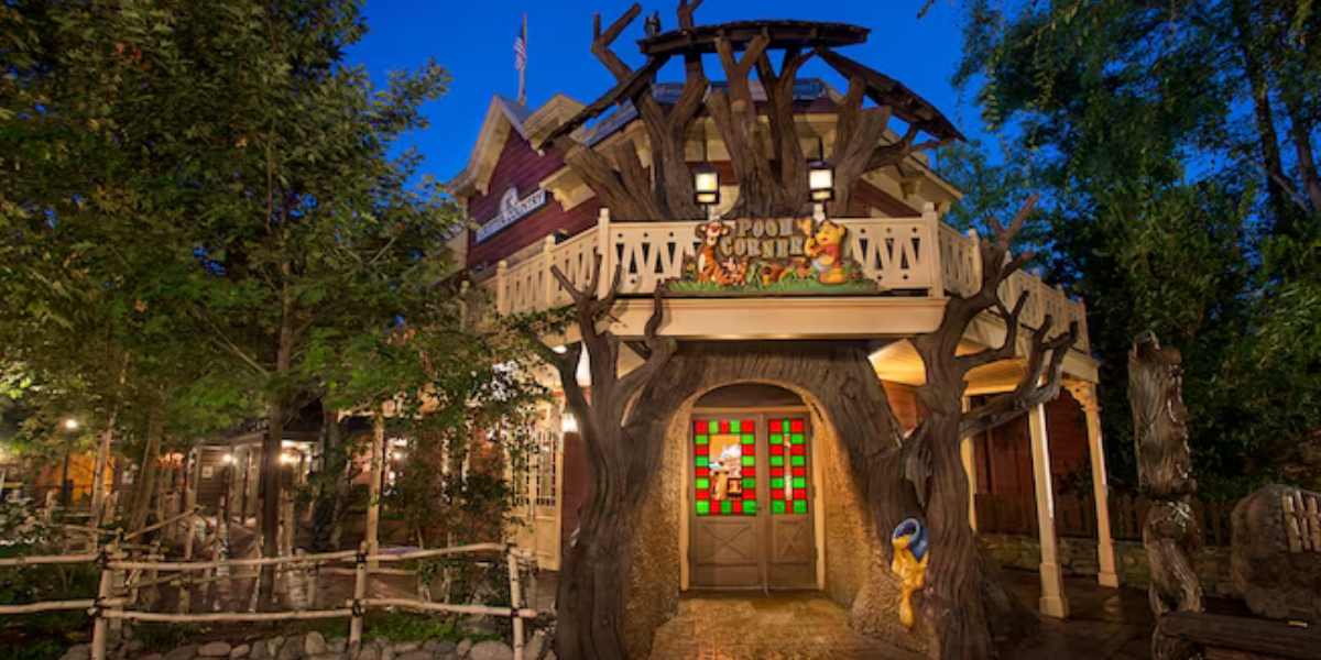 Night view of a whimsical, tree-inspired cottage with a large, colorful stained glass window and wooden sign that reads "Geppetto's Cottage," inspired by the worst Disneyland ride. It is surrounded by lush greenery and softly lit pathways.