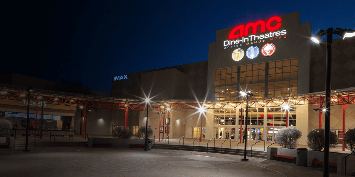 A nighttime exterior view of an AMC Dine-In Theatres location, with the illuminated AMC logo and "Dine-In Theatres" text prominently displayed above the entrance. The building also features an IMAX sign to the left. The area is well-lit with streetlights.