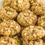 A white plate filled with a generous pile of oatmeal apple cookies. The cookies have a chunky texture with visible pieces of apple and oats, giving them a homemade, rustic appearance. The background shows part of a green apple and some foliage.