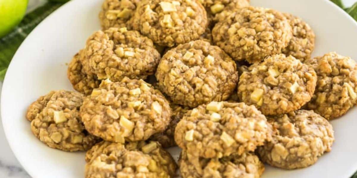 A white plate filled with a generous pile of oatmeal apple cookies. The cookies have a chunky texture with visible pieces of apple and oats, giving them a homemade, rustic appearance. The background shows part of a green apple and some foliage.