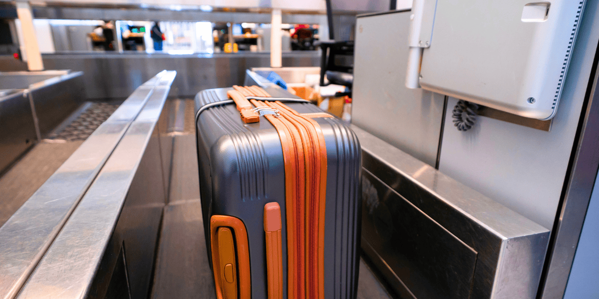 A close-up of a gray hard-shell suitcase with orange accents on a conveyor belt in an airport security checkpoint. The suitcase is positioned upright and next to other passengers' items on the conveyor belt at MCO.