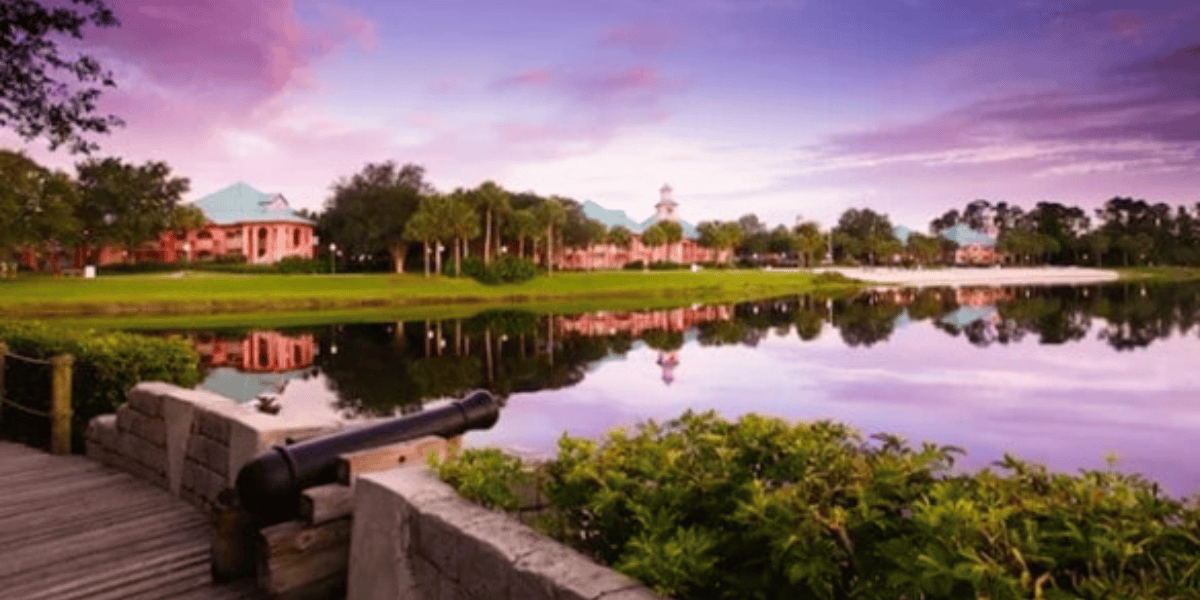 A serene lakeside scene at sunset featuring a tranquil body of water reflecting the soft pink and purple hues of the sky. A wooden boardwalk with a cannon, lush green bushes, and charming pastel-colored buildings line the opposite shore amidst trees.