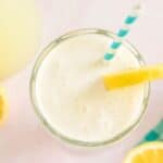 A glass of creamy lemonade with frothy top, garnished with a lemon slice and a blue-striped straw, sits on a light pink surface. Whole and halved lemons along with a blue and white checkered cloth are scattered around.