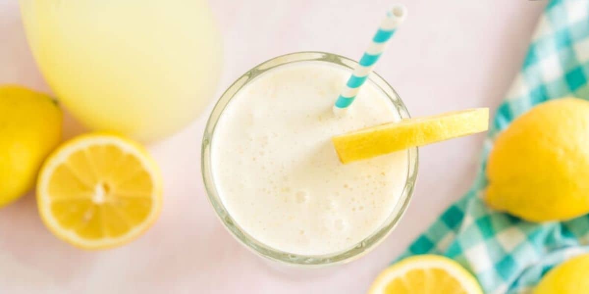 A glass of creamy lemonade with frothy top, garnished with a lemon slice and a blue-striped straw, sits on a light pink surface. Whole and halved lemons along with a blue and white checkered cloth are scattered around.