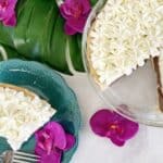 A pie garnished with white frosting and flower designs, has one slice cut out and placed on a turquoise plate with dotted edges. The setup is decorated with large green leaves and bright pink flowers, all on a white surface. A fork rests on the plate.