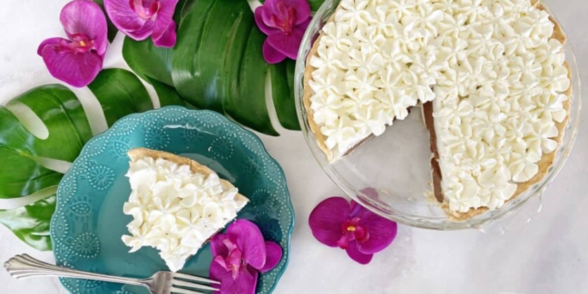 A pie garnished with white frosting and flower designs, has one slice cut out and placed on a turquoise plate with dotted edges. The setup is decorated with large green leaves and bright pink flowers, all on a white surface. A fork rests on the plate.