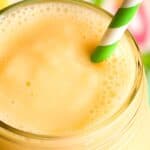 A close-up shot of a creamy yellow mango smoothie in a glass jar, topped with a green and white striped straw. The background features vibrant, colorful patterns that add a lively atmosphere.