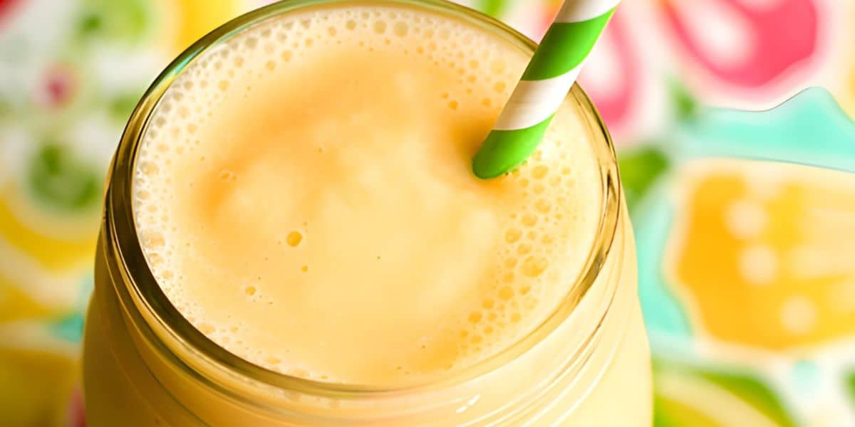A close-up shot of a creamy yellow mango smoothie in a glass jar, topped with a green and white striped straw. The background features vibrant, colorful patterns that add a lively atmosphere.