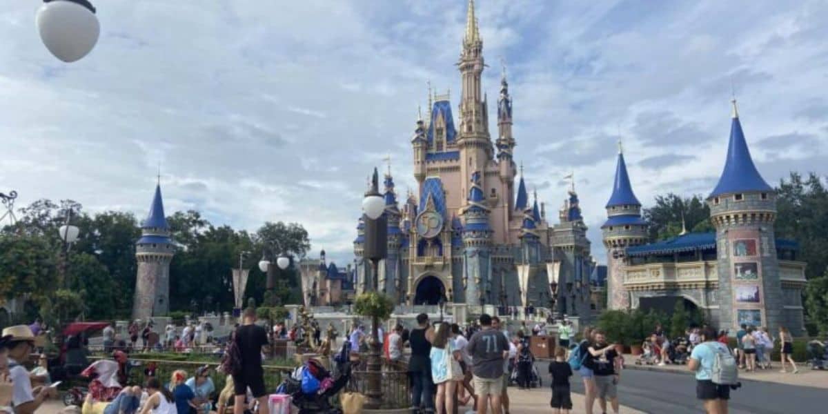 A bustling crowd of people gather in front of a large, ornate castle with blue spires at an amusement park on a sunny day. Balloons float in the air, and visitors are taking photos, strolling, and enjoying the scene. The sky is partly cloudy.