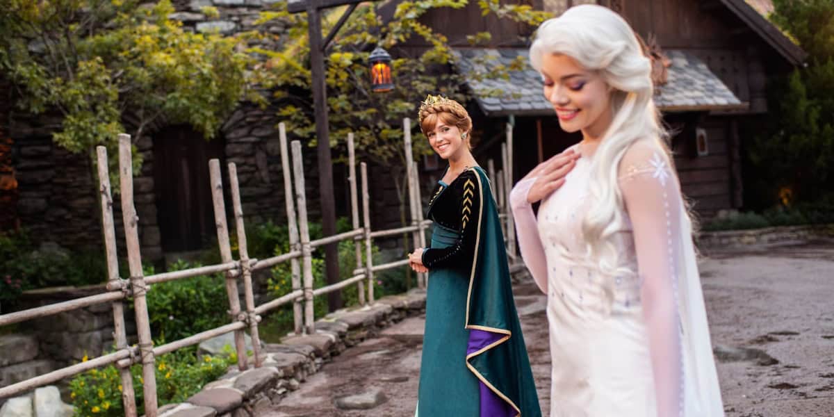 Two women dressed as princesses walk along a rustic path in an outdoor setting with stone buildings and wooden fences. One wears a teal gown and crown, and the other wears a white gown with a flowing cape. They appear to be happy and are enjoying the scenery.