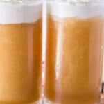 Two large glass mugs filled with a frothy, golden brown beverage topped with a layer of white foam. The mugs are placed on a wooden surface with a blurred background that includes lit candles and a red-and-white striped cloth.