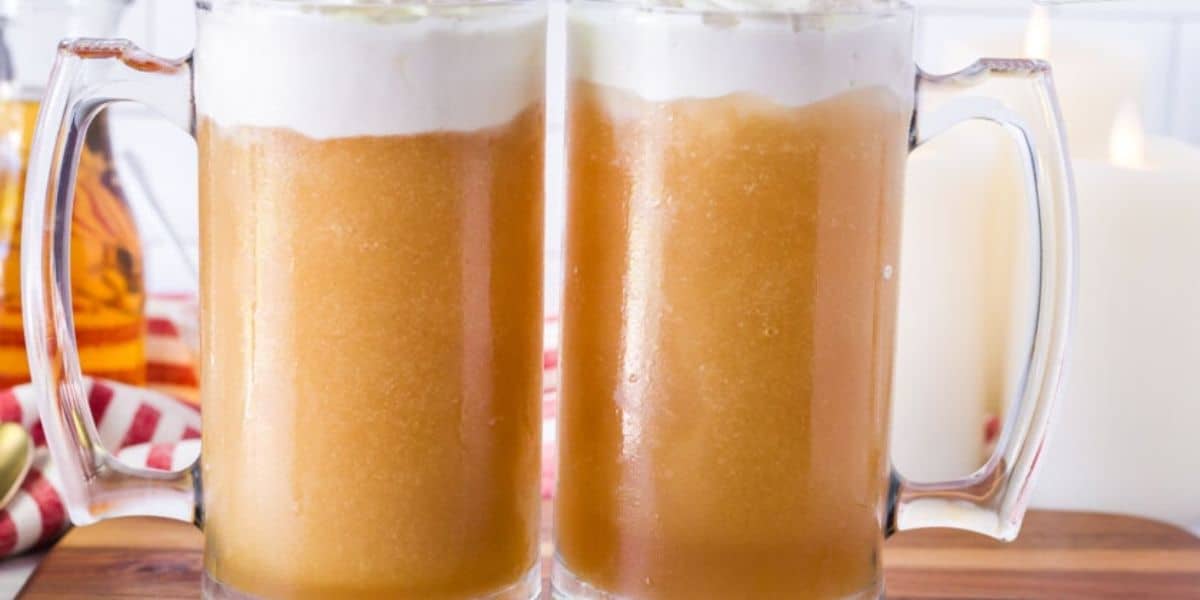 Two large glass mugs filled with a frothy, golden brown beverage topped with a layer of white foam. The mugs are placed on a wooden surface with a blurred background that includes lit candles and a red-and-white striped cloth.